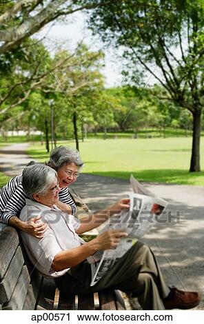 Stock Image - Couple in park, man reading newspaper, woman standing behind him looking over shoulder. Fotosearch