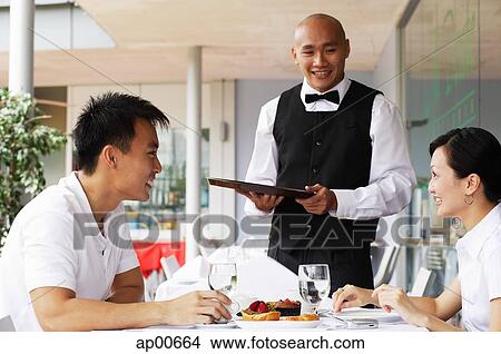 Picture - Couple in restaurant, waiter standing next to their table. Fotosearch