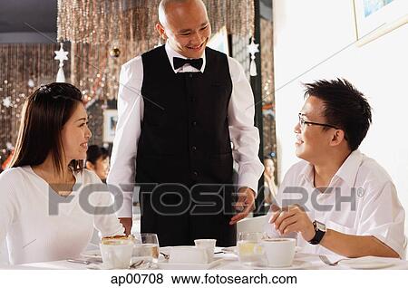 Stock Photo - Couple in restaurant, waiter standing and smiling at them. Fotosearch