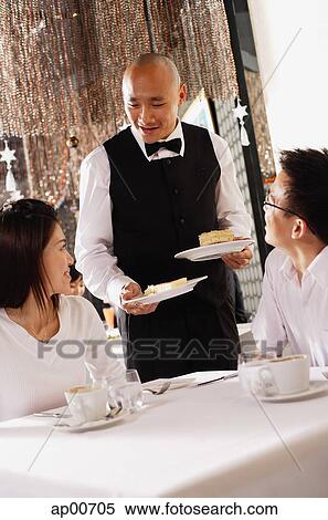 Stock Photography - Couple in restaurant, waiter standing next to their table, holding plates of dessert. Fotosearch