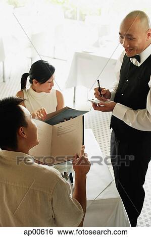 Stock Photograph - Couple looking at menus in restaurant, waiter taking their order. Fotosearch