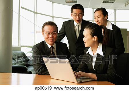 Stock Photo - Executives in office, sitting and standing around open laptop, talking. Fotosearch