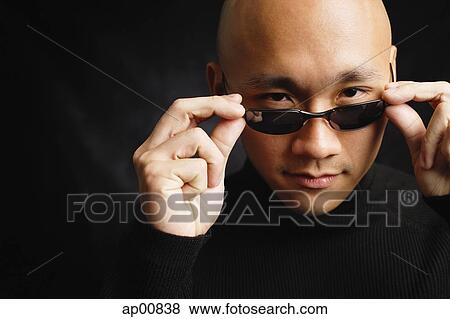 Stock Photo - Man with shaved head, adjusting sunglasses, looking at camera. Fotosearch