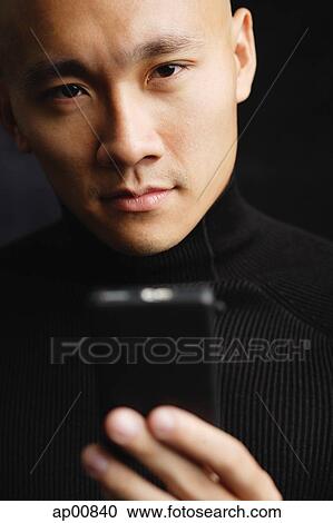 Stock Image - Man with shaved head, holding mobile phone, looking at camera selective focus. Fotosearch