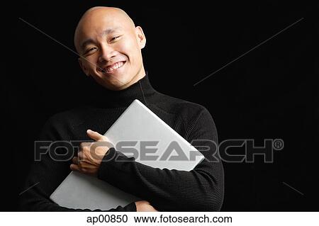 Stock Image - Man with shaved head, hugging laptop, looking at camera, smiling. Fotosearch
