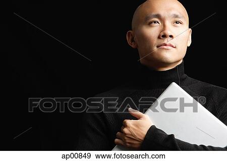 Stock Photo - Man with shaved head, hugging laptop, looking away. Fotosearch