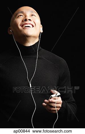Picture - Man with shaved head, listening to music with earphones, looking up smiling. Fotosearch