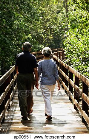 Stock Image - Mature couple walking on wooden bridge, rear view. Fotosearch