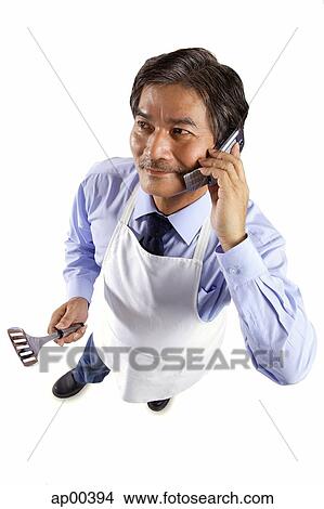 Mature man wearing apron, holding spatula and using mobile phone View Large Photo Image Picture - Mature man wearing apron, holding spatula and using mobile phone. Fotosearch