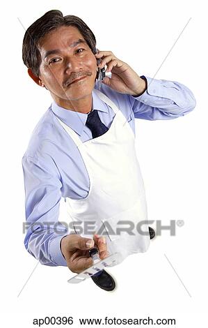 Stock Photograph - Mature man wearing apron, using mobile phone, holding spatula looking at camera. Fotosearch