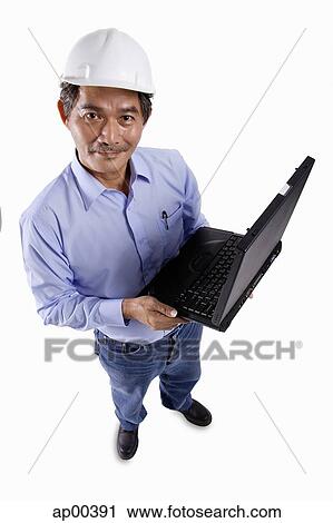 Stock Image - Mature man wearing construction hat, holding laptop, looking at camera. Fotosearch