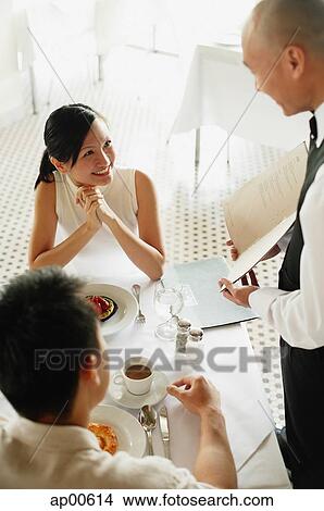 Picture - Waiter showing menu to couple at restaurant, woman smiling at him. Fotosearch