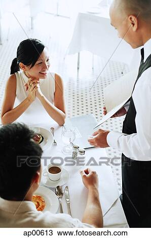 Stock Image - Waiter showing menu to couple at restaurant. Fotosearch