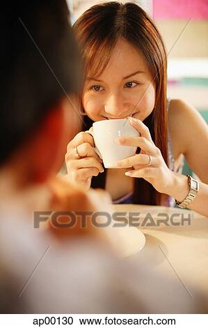 Stock Image - Woman drinking from mug, facing another person. Fotosearch
