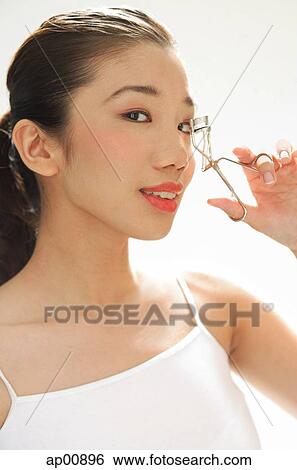 Stock Photograph - Woman holding eyelash curler to eye, looking at camera. Fotosearch