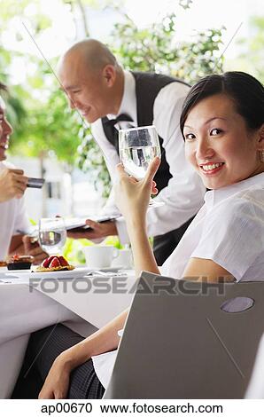 Stock Image - Woman holding glass of water, looking at camera, people in the background. Fotosearch