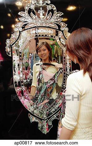 Stock Photo - Woman in clothing store, holding dress, looking at mirror. Fotosearch