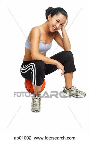 Stock Image - Woman sitting on basketball, looking at camera, smiling. Fotosearch