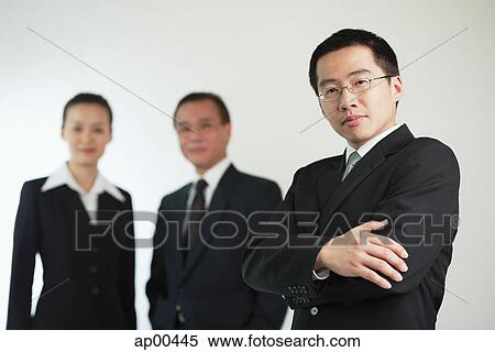 Stock Photography - Young businessman with arms crossed, businessman and businesswoman in the background. Fotosearch