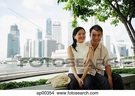 Picture - Young couple sitting on park bench, looking at camera. Fotosearch