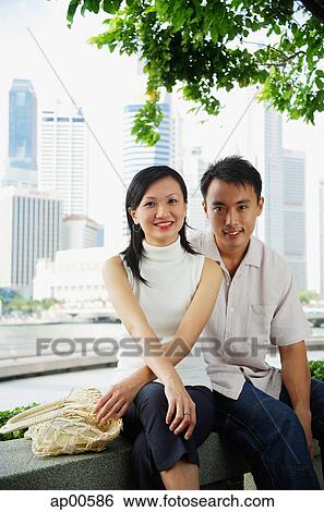 Stock Photograph - Young couple sitting on park bench, looking at camera. Fotosearch