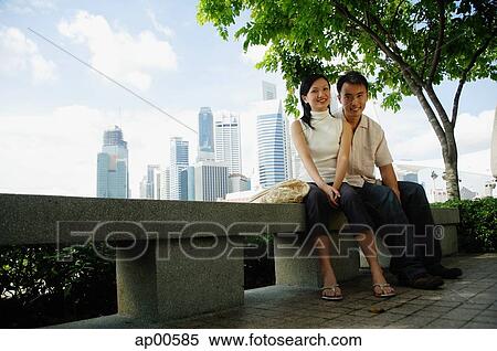 Stock Photography - Young couple sitting on park bench, looking at camera, buildings in the background. Fotosearch