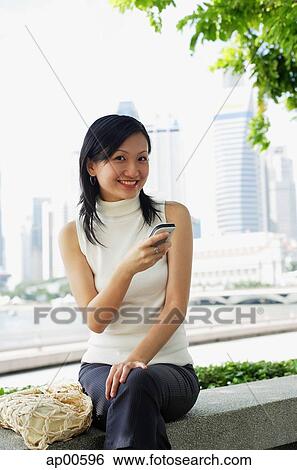 Stock Photograph - Young woman, holding mobile phone, looking at camera. Fotosearch