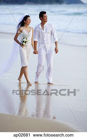 Stock Image - Bride and groom walking on beach, holding hands. Fotosearch