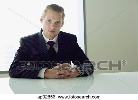 Businessman sitting at table, hands clasped, looking at camera View Large Photo Image Stock Photograph - Businessman sitting at table, hands clasped, looking at camera . Fotosearch