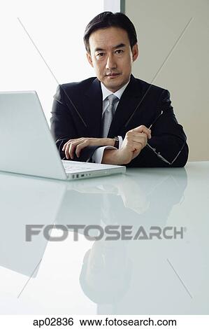 Businessman sitting at table, laptop next to him, looking at camera View Large Photo Image Stock Photograph - Businessman sitting at table, laptop next to him, looking at camera . Fotosearch