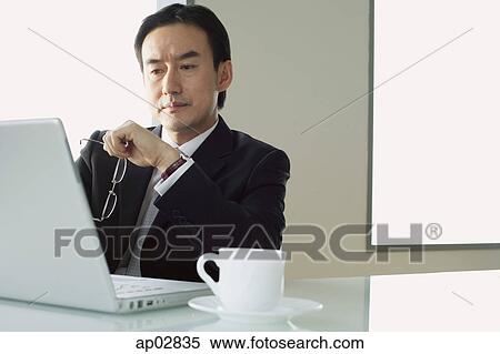 Businessman sitting at table, looking at laptop View Large Photo Image Stock Photography - Businessman sitting at table, looking at laptop . Fotosearch