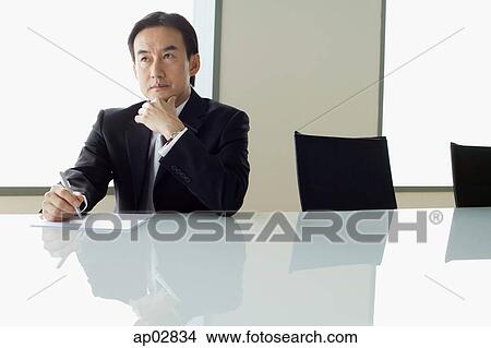 Businessman sitting at table, with pen and paper, hand on chin View Large Photo Image Picture - Businessman sitting at table, with pen and paper, hand on chin . Fotosearch