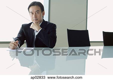 Businessman sitting at table, with pen and paper, hand on chin, looking at camera View Large Photo Image Stock Image - Businessman sitting at table, with pen and paper, hand on chin, looking at camera . Fotosearch