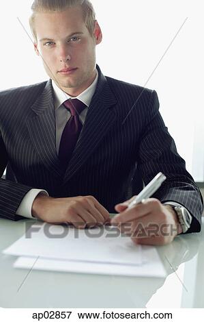 Businessman sitting at table with pen and paper, looking at camera View Large Photo Image Stock Photo - Businessman sitting at table with pen and paper, looking at camera . Fotosearch