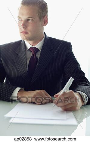 Businessman sitting at table with pen and paper, looking away View Large Photo Image Stock Photo - Businessman sitting at table with pen and paper, looking away . Fotosearch
