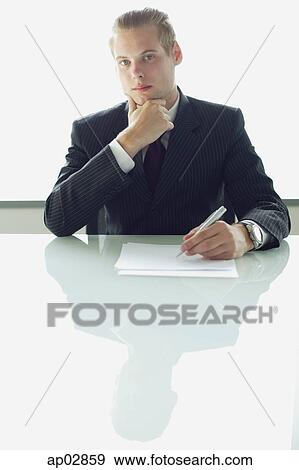 Businessman sitting at table with pen and paper, looking at camera, hand on chin View Large Photo Image Stock Photo - Businessman sitting at table with pen and paper, looking at camera, hand on chin . Fotosearch