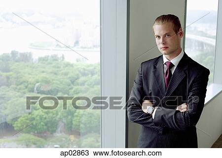Businessman standing next to window in conference room, arms crossed, looking at camera View Large Photo Image Stock Image - Businessman standing next to window in conference room, arms crossed, looking at camera . Fotosearch