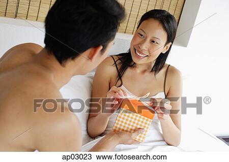 Stock Image - Couple in bed, man holding gift box, woman unwrapping ribbon . Fotosearch