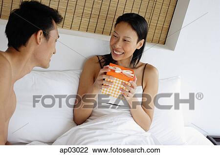 Stock Image - Couple in bed, woman holding gift box, smiling . Fotosearch