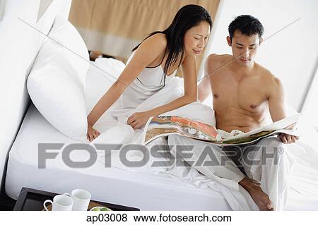 Stock Photo - Couple sitting on bed, looking at newspaper. Fotosearch