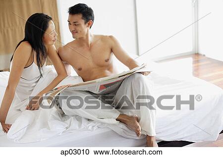 Stock Image - Couple sitting on bed with newspaper, looking at each other . Fotosearch