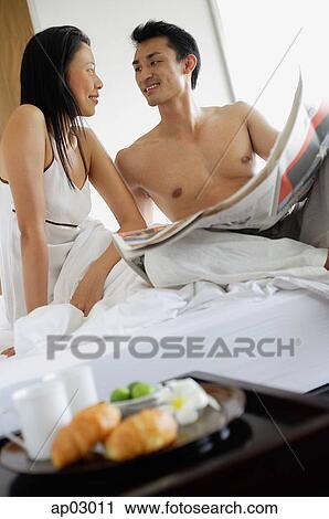 Couple sitting on bed with newspaper, looking at each other, smiling View Large Photo Image Stock Image - Couple sitting on bed with newspaper, looking at each other, smiling . Fotosearch