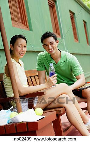 Stock Photo - Couple sitting on bench on tennis court, smiling at camera, woman holding water bottle . Fotosearch