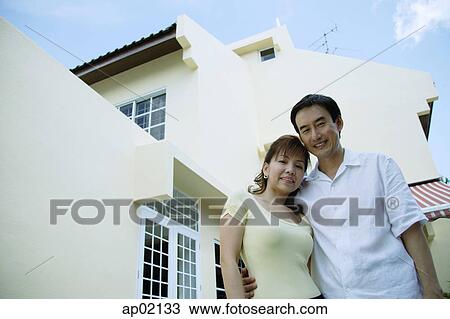 Stock Image - Couple standing in front of house, arms around each other . Fotosearch