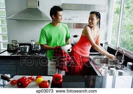 Stock Image - Couple standing in kitchen, woman washing hands, man drying dishes . Fotosearch