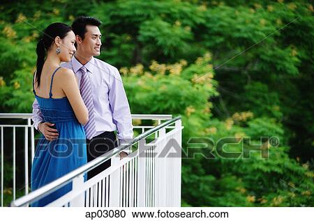 Stock Image - Couple standing on balcony, looking away, side view. Fotosearch
