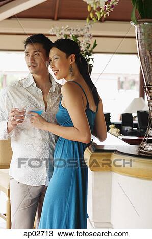 Stock Image - Couple standing side by side, holding drinks, looking away . Fotosearch