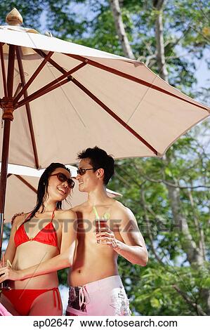 Stock Photo - Couple standing under umbrella, holding drinks, looking at each other . Fotosearch