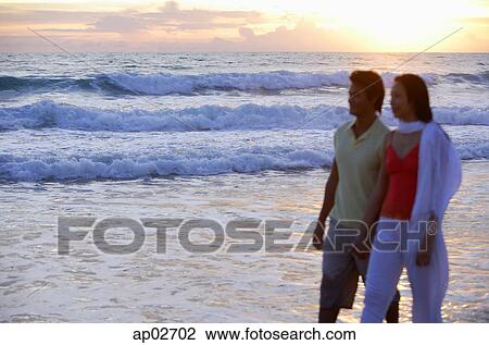 Stock Image - Couple walking on beach, during sunset, holding hands . Fotosearch