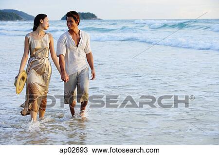 Stock Image - Couple walking on beach, holding hands, looking at each other . Fotosearch